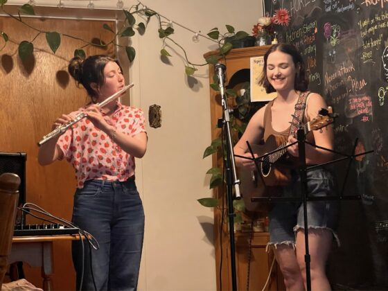 Two people, one playing the flute and another playing guitar, perform in a living room.